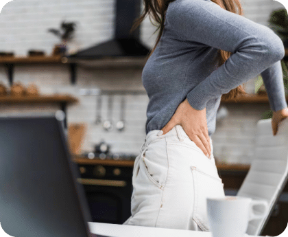 Woman standing in a kitchen at a laptop holding her lower back in pain